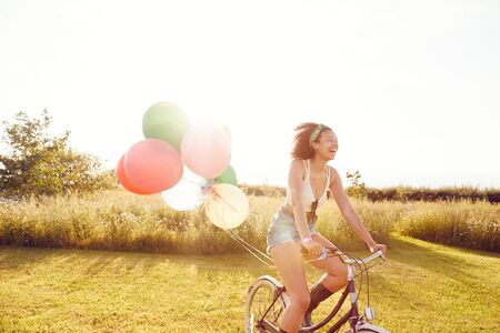 Young Woman Riding Bicycle Decorated With Balloons Through Countryside Against Flaring Sunの写真素材