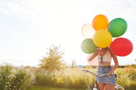 Young Woman With Bicycle Hidden By Balloons Rides Through Countryside Against Flaring Sunの写真素材