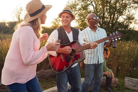 Group Of Mature Friends Sitting Around Fire As They Drink And Sing Songs At Outdoor Campsiteの写真素材