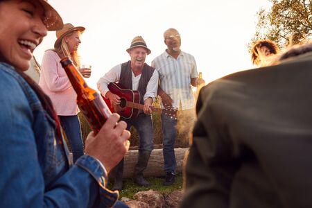 Group Of Mature Friends Sitting Around Fire As They Drink And Sing Songs At Outdoor Campsite
