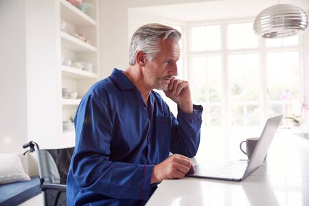 Mature Disabled Man In Wheelchair At Home Using Laptop On Kitchen Counter