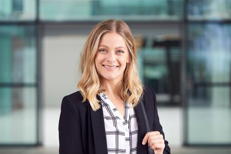 Head And Shoulders Portrait Of Smiling Businesswoman Standing In Lobby Of Busy Modern Office