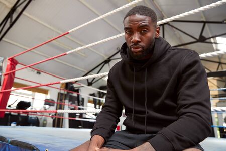 Portrait Of Male Boxer Wearing Hooded Sweatshirt In Gym Sitting On Boxing Ringの写真素材