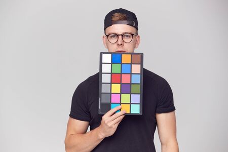 Portrait Of Male Photographer In Studio Holding Colour Checker Card Against White Background