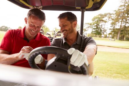 Two Mature Men Playing Golf Marking Scorecard In Buggy Driving Along Courseの写真素材