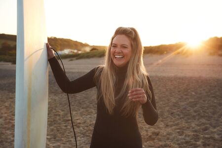 Woman Wearing Wetsuit Holding Surfboard Enjoying Surfing Staycation On Beach As Sun Setsの写真素材