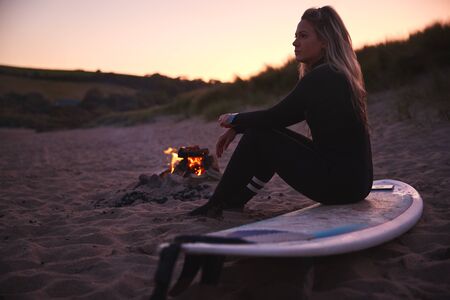 Woman Sitting On Surfboard By Camp Fire On Beach As Sun Sets Behind Herの写真素材