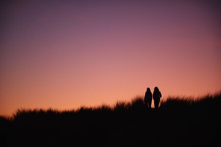 Silhouette Of Two Female Friends Walking Across Dunes Against Setting Sunの写真素材