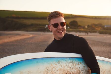 Young Man In Sunglasses Wearing Wetsuit Enjoying Surfing Staycation On Beach As Sun Setsの写真素材