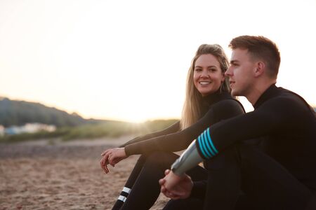 Profile View Of Couple Wearing Wetsuits On Surfing Staycation Sitting On Beach Looking Out To  Seaの写真素材
