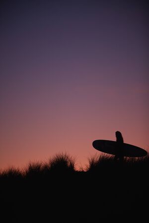 Silhouette Of Female Surfer Carrying Surfboard Across Dunes Against Setting Sunの写真素材