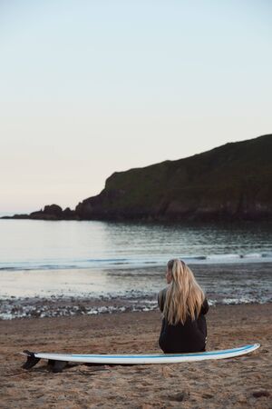 Rear View Of Thoughtful Woman Wearing Wetsuit On Surfing Staycation Looking Out To  Sea At Waves