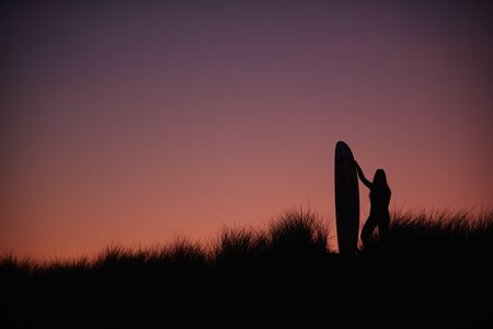 Silhouette Of Female Surfer Standing And Holding Surfboard Across Dunes Against Setting Sunの写真素材