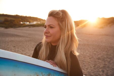 Woman Wearing Wetsuit Holding Surfboard Enjoying Surfing Staycation On Beach As Sun Sets