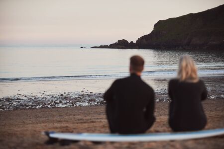 Defocused Shot Of Couple In Wetsuits On Surfing Staycation Sitting On Surfboard Looking Out To  Sea