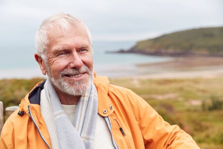 Active Senior Man Walking Along Coastal Path In Fall Or Winter By Gate With Beach And Cliffs Behind