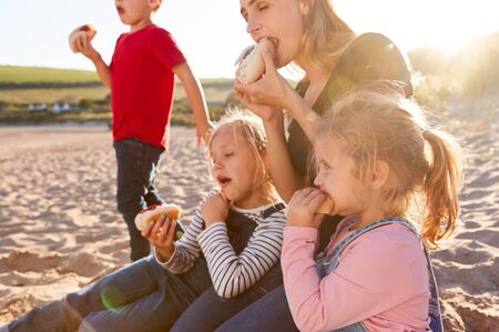 Mother With Children Eating Hot Dogs Sitting On Sand At Beach Barbecueの写真素材