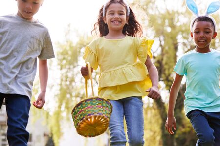 Group Of Children Wearing Bunny Ears Running On Easter Egg Hunt In Garden