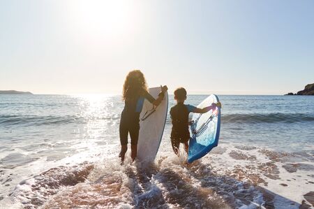 Rear View Of Two Children Wearing Wetsuits Running Into Sea With Bodyboards On Beach Vacationの写真素材