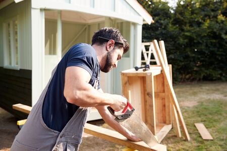 Male Carpenter With Female Apprentice Sawing Wood To Build Outdoor Summerhouse In Gardenの写真素材