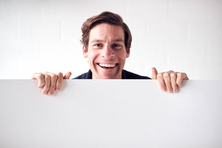 Portrait Of Happy Young Man Holding Blank White Card With Copy Space Standing Against White Wallの写真素材