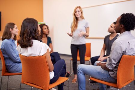 Woman Standing To Address Group Of Men And Women At Mental Health Group Therapy Meeting