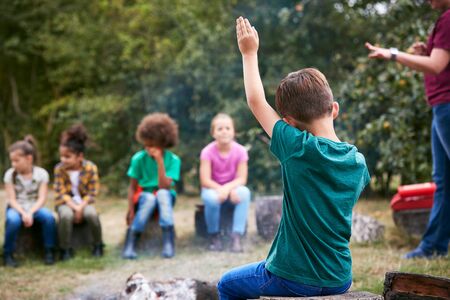 Children On Outdoor Activity Camping Trip Sit Around Camp Fire With Arms Raised Answering Questionの写真素材