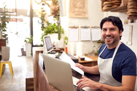 Portrait Of Male Sales Assistant Working On Laptop Behind Sales Desk Of Florists Storeの写真素材