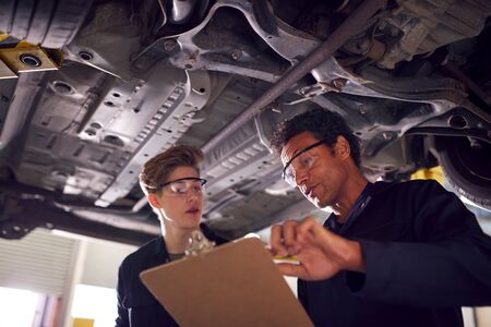 Male Tutor With Student Looking Underneath Car On Hydraulic Ramp On Auto Mechanic Courseの写真素材