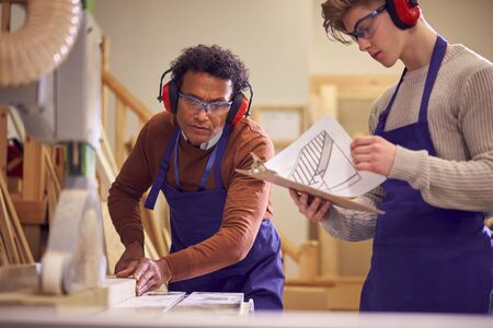 Tutor With Male Carpentry Student In Workshop Studying For Apprenticeship At College Using Bench Saw