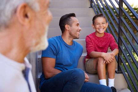 Smiling Multi-Generation Male Hispanic Family Sitting On Steps In Garden And Talking Together
