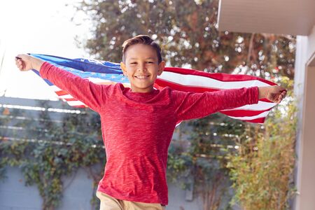 Portrait Of Proud Hispanic Boy Wrapped In Stars And Stripes American Flag Running Towards Cameraの写真素材