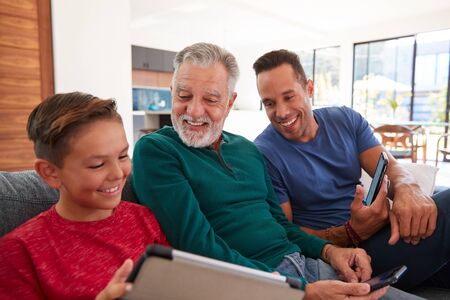 Multi-Generation Male Hispanic Family Sitting On Sofa At Home Using Mobile Phones And Digital Tabletの写真素材