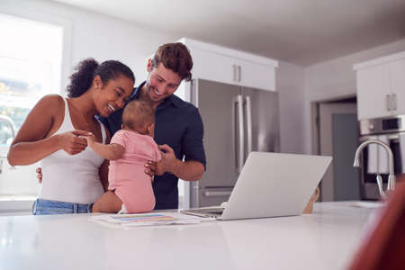 Family With Baby Daughter In Kitchen Using Laptop On Counter