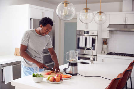 Man Wearing Pyjamas Standing In Kitchen Chopping Fruit And Vegetables For Fresh Smoothieの写真素材