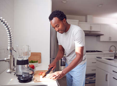 Man Wearing Pyjamas Standing In Kitchen Chopping Fruit For Fresh Smoothieの写真素材