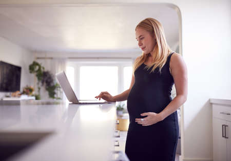 Pregnant Woman Standing By Kitchen Counter Working From Home On Laptopの写真素材