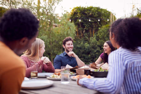 Multi-Cultural Friends At Home Sitting At Table Enjoying Food At Summer Garden Partyの写真素材