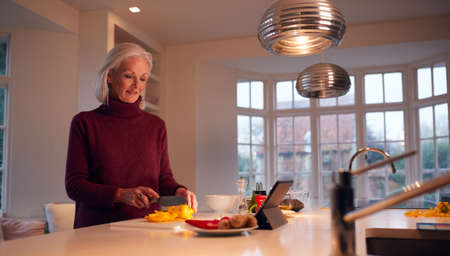 Senior Woman In Kitchen At Home Following Recipe On Digital Tablet