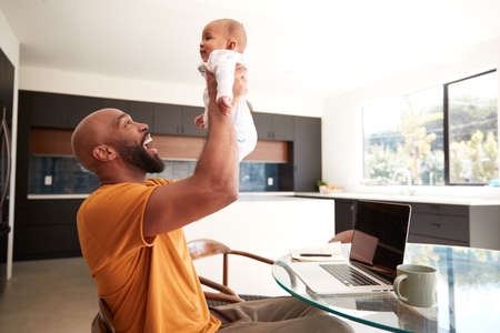 Smiling Stay At Home African American Father Cuddling Baby Daughter Whilst Working On Laptop At Homeの写真素材