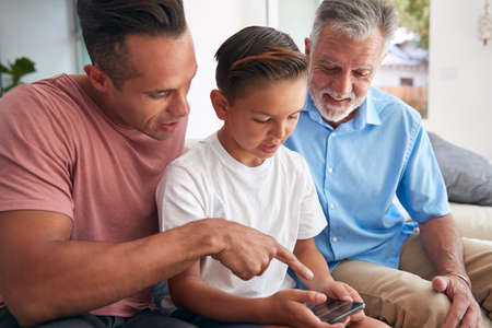 Multi-Generation Male Hispanic Family  Using Mobile Phone On Sofa At Home Togetherの写真素材