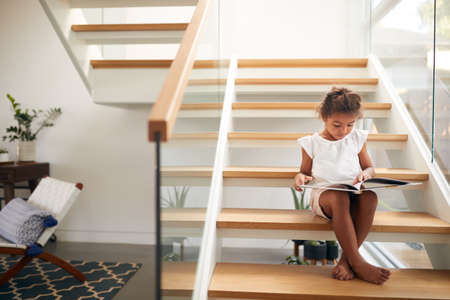 Hispanic Girl Sitting On Staircase In Modern Home Reading Bookの写真素材
