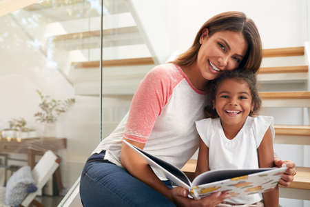 Portrait Of Hispanic Mother And Daughter Sitting On Staircase In Modern Home Reading Book Together