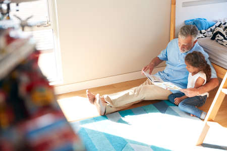 Hispanic Grandfather And Granddaughter Sitting On Floor Of Childrens Bedroom Reading Book Togetherの写真素材