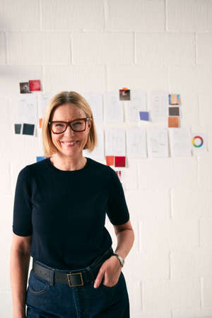 Portrait Of Mature Woman Standing In Front Of Designs On Wall In Start Up Fashion Businessの写真素材