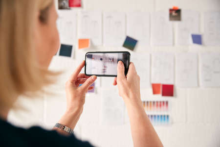 Female Designer Taking Photos Of Designs On Wall With Mobile Phone In Start Up Fashion Businessの写真素材