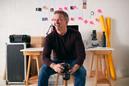 Laughing Mature Male Photographer With Camera Sitting On Stool In Front Of Desk In Studioの写真素材