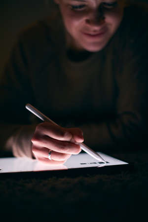 Close Up Of Woman Drawing On Digital Tablet Using Stylus Pen Lying On Carpet At Night