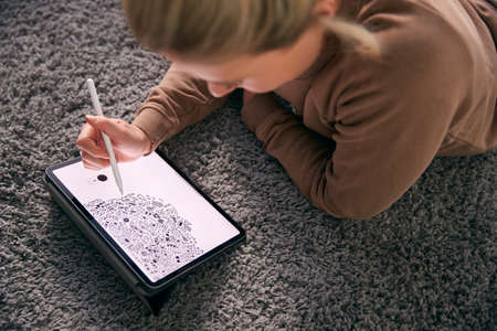 Close Up Of Woman Drawing On Digital Tablet Using Stylus Pen Lying On Carpet At Homeの写真素材