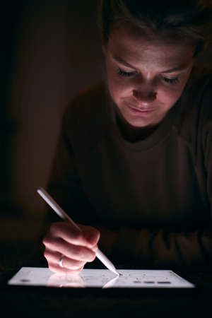 Close Up Of Woman Drawing On Digital Tablet Using Stylus Pen Lying On Carpet At Nightの写真素材
